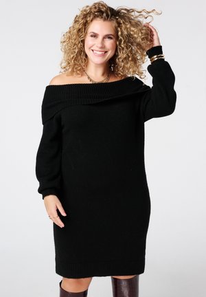 Zwarte off-shoulder gebreide jurk met lange mouwen, textuur met ribbelige halslijn en manchetten. Model gestyled met gouden accessoires en donkerbruine laarzen.