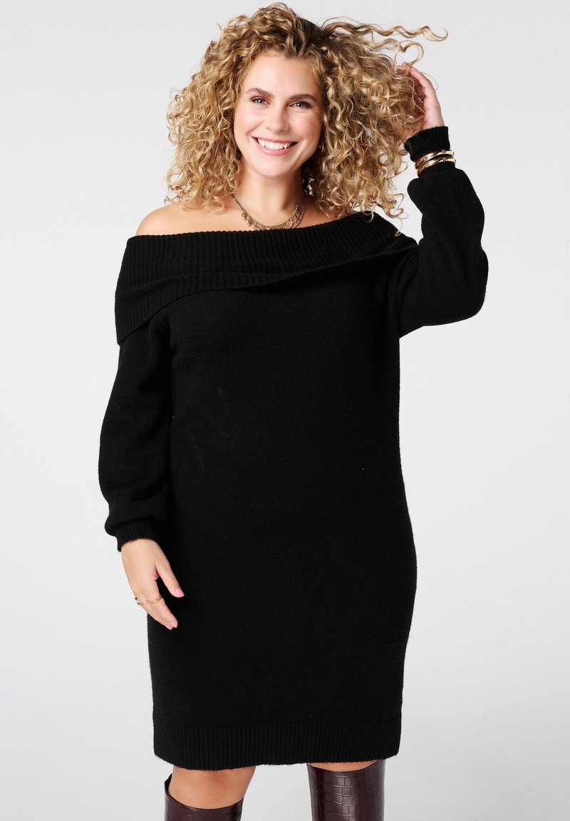 Zwarte off-shoulder gebreide jurk met lange mouwen, textuur met ribbelige halslijn en manchetten. Model gestyled met gouden accessoires en donkerbruine laarzen.