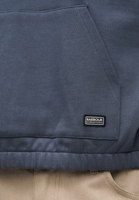 Σκούρο μπλε φούτερ Barbour International με τσέπη και λογότυπο πάνω από τη μπεζ μέση του παντελονιού.
