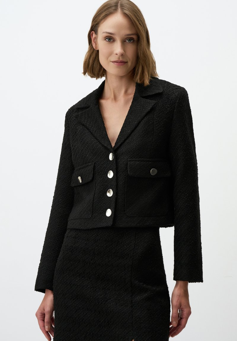 Schwarze (kurz geschnittene) Jacke aus strukturiertem Stoff mit Spitzkragen, zwei vorderen Taschen und fünf großen silberfarbenen Knöpfen.