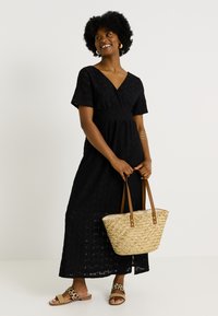 Femme souriante, portant une robe midi noire texturée, des sandales imprimé léopard, des boucles d'oreilles et des bracelets en or, tenant un sac fourre-tout en paille tissée.