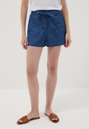 Mujer con pantalones cortos de denim azul con bolsillos delanteros y cinturón de tela, combinados con sandalias planas marrones y una camiseta blanca.