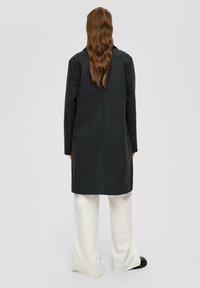 Langer dunkelgrauer Mantel aus strukturiertem Stoff, mit geradem Schnitt, Reverskragen und Nahtdetails hinten, getragen über weißen weit geschnittenen Hosen.