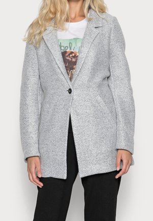 Kvinde iført en lysegrå uldfrakke med enkelt knap, hvid grafisk t-shirt med hundeface og sorte bukser, stående mod en ensfarvet baggrund.