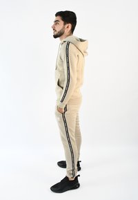 Sudadera beige y pantalones de chándal a juego, confeccionados en tejido suave, con franjas laterales negras con logo, combinados con zapatillas deportivas negras.