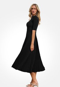 Zwart geribbelde midi-jurk met een nauwsluitend lijfje en een uitlopende geplooide rok. Korte mouwen en een gladde textuur. Gemaakt met lichte gekleurde hoge hakken.
