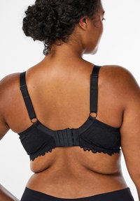 Reggiseno in pizzo nero con spalline regolabili, chiusura posteriore a sei ganci e bordi in pizzo ondulato. Il tessuto presenta un motivo floreale a rilievo.