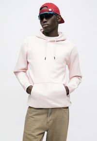 Rosa Kapuzenpullover aus weichem Material mit einer Kängurutasche, kombiniert mit hellbraunen Hosen. Mit roter Mütze und dunkler Brille ausgestattet.