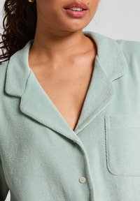 Vrouw in een lichtgroene badstof knoopshirt met zak, gouden oorring en gedeeltelijk zichtbare krullende haren die naar achteren zijn gebonden.