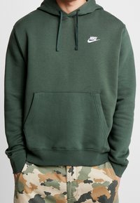 Grön Nike-huvtröja med ficka fram, snörning i huvan och liten vit logotyp på vänster bröstområde. Mjuk tygtextur.