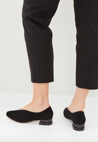 Czarne zamszowe buty na niskim obcasie z okrągłym noskiem i wyciętym wzorem. Posiadają geometryczny, kontrastowy obcas i gładką fakturę.