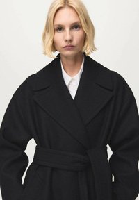 Zwarte wollen jas met brede revers en een ceintuur in de taille. Heeft een gestructureerde stof en zijzakken. Eronder een strak wit overhemd.