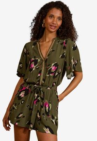 Olīvu zaļš romperis ar īsām piedurknēm, V veida kakla izgriezumu, zelta apdari un auklu viduklī. Satur leoparda rakstu ar rozā un melniem akcentiem.