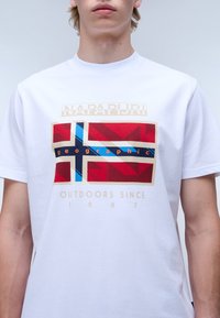 Bílý bavlněný tričko s grafikou norské vlajky v červené, modré a zlaté barvě, s textem "NAPAPIJRI" a "geographic".