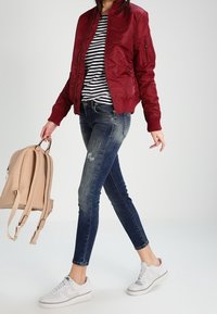 Burgundská bomber bunda přes pruhované tričko, spárovaná s roztrhanými modrými džíny a bílými teniskami; nese béžový batoh.