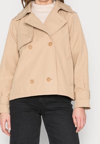 Beige double-breasted trench coat with large buttons, side pockets, and a wide collar; made of smooth, lightweight fabric.