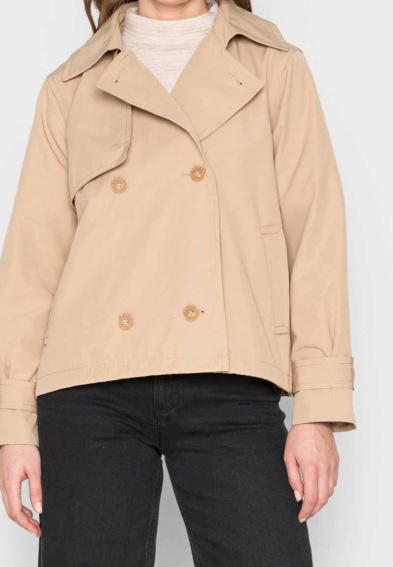Beige double-breasted trench coat with large buttons, side pockets, and a wide collar; made of smooth, lightweight fabric.