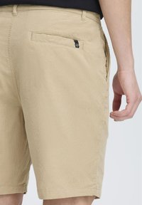 Shorts beiges en tissu texturé, dotés d'une poche arrière avec un petit logo noir et d'une coupe décontractée, se terminant juste au-dessus du genou.
