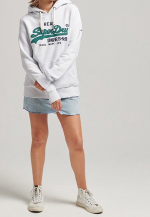 Frau trägt hellgrauen Superdry-Hoodie, hellblauen Jeans-Minirock und weiße High-Top-Sneaker, steht vor schlichtem Hintergrund.