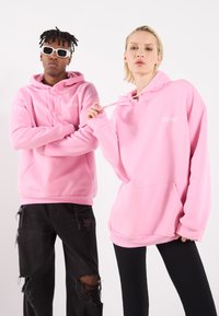 Felpe rosa con tasche frontali e cordini, con "LATE NIGHT" ricamato in bianco. Un modello indossa occhiali da sole, abbinati a jeans neri strappati.
