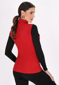 Rotes ärmelloses Oberteil mit hohem Kragen, kombiniert mit einem schwarzen Langarmshirt darunter, aus glattem Stoff und mit taillierter Silhouette.