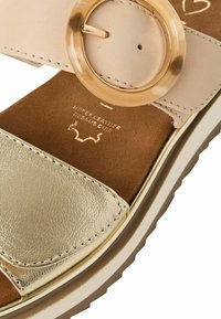 Sandale beige clair avec une lanière en cuir doré et une grande boucle ronde en acrylique. Semelle intérieure marron douce et semelle épaisse blanche.