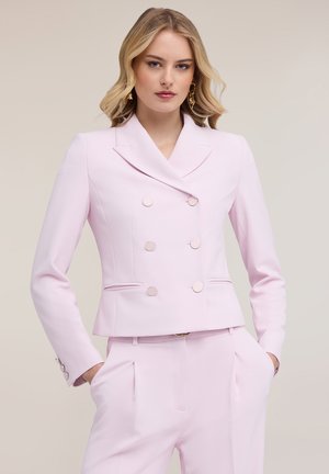 Blonde Frau trägt ein hellrosa doppelreihiges Sakko und passende Hosen, die Hände in den Taschen, vor einem neutralen Hintergrund.