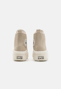 Beżowe sneakersy typu high-top z teksturowanym materiałem, białą gumową podeszwą i okrągłą naszywką logo na zewnętrznej części kostki.