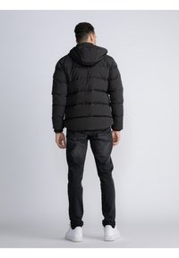 Chaqueta tipo puffer negra con capucha, con un diseño acolchado y puños elásticos. Llevada sobre vaqueros oscuros y zapatillas blancas, vista desde atrás.