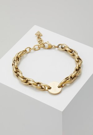 Bracelet chaîne en or avec des maillons de taille moyenne, un pendentif rond gravé "GUESS" et un fermoir mousqueton sur un socle blanc.