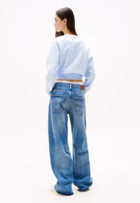 Žena s dlhými tmavými vlasmi, oblečená v svetlomodrej krátkej mikine a voľných modrých džínsach, stojaca a pozerajúca sa do strany na bielom pozadí.