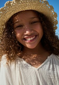 Chapeau de paille avec un tissage texturé, haut beige clair avec un motif subtil, et un visage souriant avec des cheveux bouclés éclairés par une douce lumière solaire.