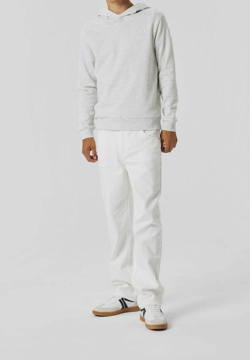 Personne portant un sweat à capuche gris clair, un pantalon blanc et des baskets blanches à rayures noires, debout sur un fond clair uni.