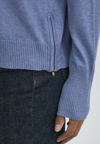 Pull en tricot bleu clair avec des poignets et un ourlet côtelés, présentant un détail de fermeture éclair sur le côté. Porté sur un jean en denim noir.