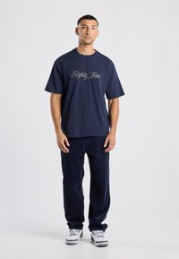 Camiseta azul marino con gran estampado de "Eighty Nine", combinada con pantalones de pana oscuros y zapatillas blancas. Estilo casual, corte relajado.
