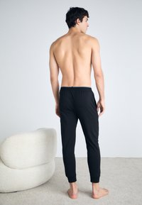 Pantalons de lounge noirs, ajustés, avec taille élastique et jambes fuselées ; texture lisse. Le modèle est pieds nus et fait légèrement face vers la droite.