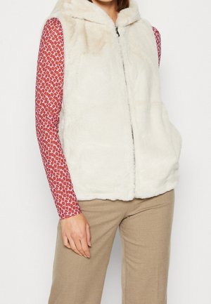 Vrouw draagt een witte pluizige mouwloze hoodie-vest over een rood bloesje met lange mouwen en beige broek, staand met een hand ontspannen.
