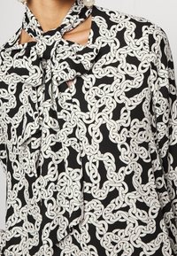 Blusa negra con un patrón de cadenas blancas, con un cuello anudado y detalles cortados en los hombros, fabricada con una tela suave.