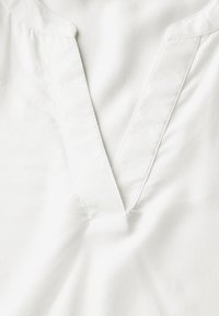 Blusa bianca in cotone con scollatura a V, tessuto morbido e cuciture minime lungo i bordi. Senza fantasie o decorazioni.