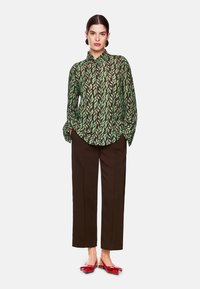 Grüne Bluse mit langen Ärmeln und schwarzem Blättermuster, kombiniert mit braunen geraden Hosen und roten Ballerinas mit Schleifendetail. Glatte Stoffoberfläche.