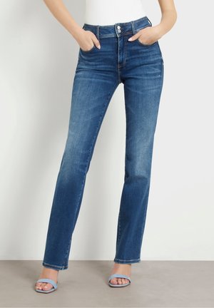 Femme portant un jean bleu taille haute, jambes droites, les mains dans les poches et des talons ouverts bleu clair sur un fond neutre.