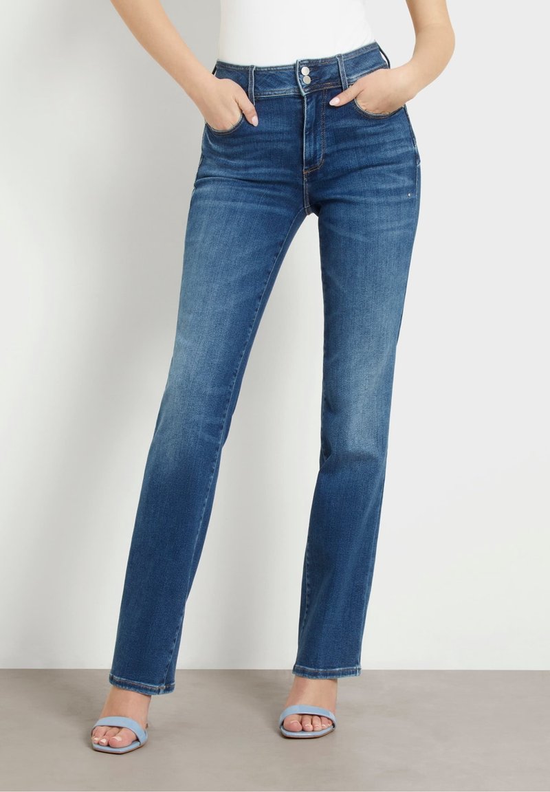 Femme portant un jean bleu taille haute, jambes droites, les mains dans les poches et des talons ouverts bleu clair sur un fond neutre.
