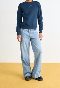 Námořnická modrá mikina s bílým logem Nike, kombinována s světle modrými širokými džíny a černými platformovými botami na žluté podlaze.