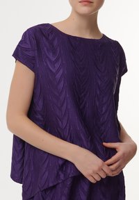 Paarse top met een textuurpatroon van verhoogde blaadvormen. Korte mouwen en een bootnek, gemaakt van een gladde, glanzende stof.