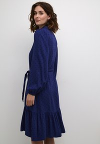 Tumesinine kleit mustade täppidega, pikad skrõsad, seotud vöökoht ja rippuv hem, valmistatud kergest kangast. Elegantne ja lihtne disain.