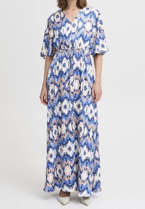 Vrouw draagt een lange blauw-wit-beige jurk met patroon, een V-hals, korte mouwen en een tailleriem, gecombineerd met witte puntige schoenen.