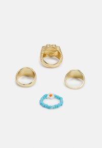 Quatro anéis: duas alianças de ouro, um anel de ouro texturizado com um design em forma de garra e um anel com contas com elementos nas cores azul, branco e laranja.