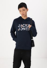 Marineblaue Kapuzen sweatshirt mit einer Kängurutasche, die das Logo "JACK & JONES" in gemustertem Text zeigt. Das Modell trägt schwarze Hose und hält eine Tasche.