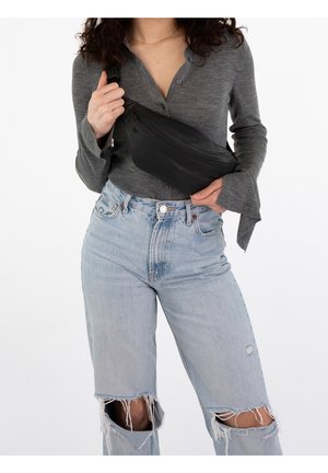 Femme portant un haut gris à boutons et un jean bleu clair déchiré, tenant un sac bandoulière noir croisé sur la poitrine.