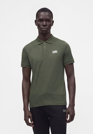 Homme portant un polo vert foncé à manches courtes et un pantalon noir, tous deux avec des logos blancs "Karl Lagerfeld", debout devant un fond blanc uni.